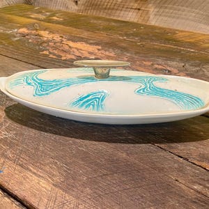 May include: Oval-shaped, white ceramic serving dish with a lid. The lid features a turquoise, abstract wave pattern and a small, clear handle. The dish is set on a weathered, wooden surface, suggesting a vintage or antique aesthetic.