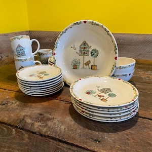 May include: A set of white ceramic dishes with a green and brown floral design. The dishes feature a birdhouse with a red heart and a bird on a branch. The dishes are arranged on a wooden table.