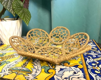 May include: A decorative, open-weave bowl in a natural tan colour. The bowl has a unique, organic shape with a series of connected circular elements. The bowl is made of a woven material, possibly jute or a similar fibre. The bowl is displayed on a colourful, patterned surface.
