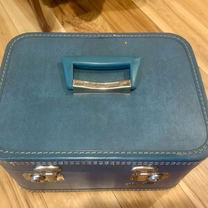 May include: A rectangular blue train case with a textured finish, featuring a matching blue plastic handle with a silver-tone metal accent, white contrast stitching along the edges, and two silver-tone metal latches on the front panel.