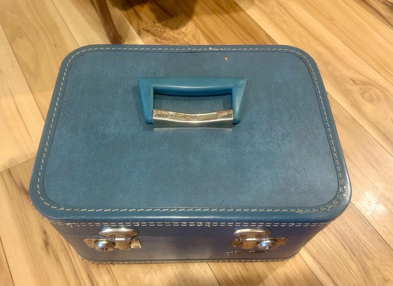 May include: A rectangular blue train case with a textured finish, featuring a matching blue plastic handle with a silver-tone metal accent, white contrast stitching along the edges, and two silver-tone metal latches on the front panel.