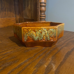 May include: A hexagonal brown ceramic planter with a green glaze and a leafy design. The planter is sitting on a wooden surface.