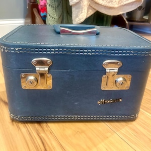 May include: A rectangular blue hard-shell travel case with a top handle and silver-toned metal hardware. The case features two front locking latches and the word Monarch in gold-toned lettering on the lower right side with white stitching.