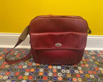 May include: A burgundy leather shoulder bag with a flap pocket and a silver-colored clasp. The bag has a long strap and is set against a yellow wall and a patterned floor.