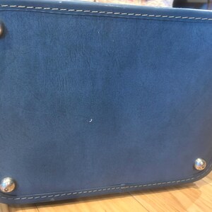 May include: A rectangular blue leather bag base featuring rounded corners and white contrast stitching along the perimeter. Four polished silver-tone metal feet are attached to the corners of the bottom panel to provide stability and protection.