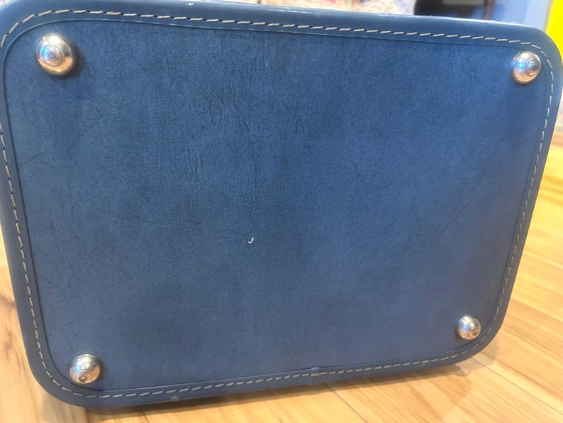 May include: A rectangular blue leather bag base featuring rounded corners and white contrast stitching along the perimeter. Four polished silver-tone metal feet are attached to the corners of the bottom panel to provide stability and protection.