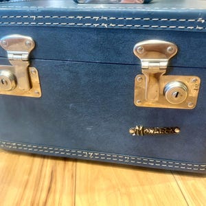May include: A rectangular blue hard-shell travel case featuring white contrast stitching along the top and bottom edges. The front includes two metallic gold-toned locking latches and a small gold-toned plaque with the text Monarch.