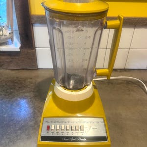Peut inclure: Un mixeur jaune à sept vitesses avec un pichet en plastique transparent doté de graduations en tasses et en onces. La base présente un panneau de commande argenté avec un bouton rouge et six boutons blancs indiquant les vitesses et fonctions.