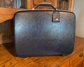 May include: A vintage black leather suitcase with a silver handle and a luggage tag. The suitcase has a textured surface and is sitting on a wooden surface.