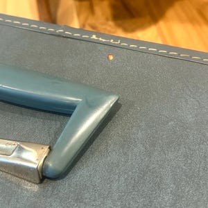 May include: A blue suitcase featuring a smooth, matte-textured surface with visible light-colored stitching along the top edge. The suitcase includes a molded blue plastic handle attached to a metallic silver-toned hinge mechanism.