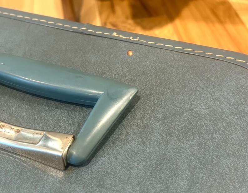 May include: A blue suitcase featuring a smooth, matte-textured surface with visible light-colored stitching along the top edge. The suitcase includes a molded blue plastic handle attached to a metallic silver-toned hinge mechanism.