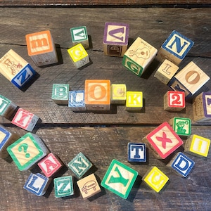May include: A collection of colorful wooden alphabet blocks scattered on a rustic wooden surface. The blocks feature letters, numbers, and simple illustrations in various colors, including red, blue, yellow, and green. The blocks are a classic educational toy.