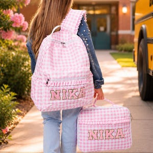 May include: A pink and white gingham backpack and matching lunch bag, both personalized with the name "NIKA" in a decorative font. The backpack has ruffled trim and is carried by a person walking towards a school building.