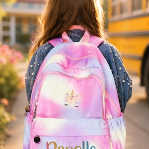 May include: A pink, purple, and white backpack with a unicorn design and the name "Danella" in glitter lettering. The backpack has a front pocket, side pocket, and adjustable straps. The image shows the back of a person wearing the backpack.