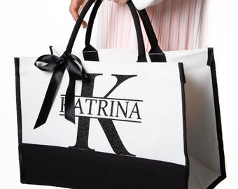 Könnte beinhalten: Weiße Tragetasche mit schwarzen Akzenten und Griffen, gehalten von einer Person. Die Tasche trägt den Namen "Katrina" mit einem großen, glitzernden "K". Eine schwarze Schleife ist an der Seite gebunden.