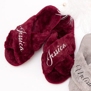 May include: Plush burgundy and gray cross-band slippers with embroidered names. The slippers are arranged on a white surface with decorative pearls and feathers. The names "Jessica", "Amelia", and "Melissa" are embroidered in white.