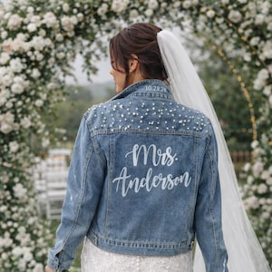 May include: A light blue denim jacket with pearl embellishments and the name "Mrs. Anderson" in white script. The jacket is worn over a white lace wedding dress. The date "10.28.21" is above the name.