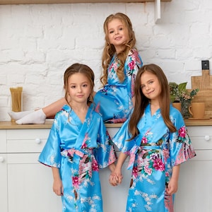 May include: Three girls wearing matching light blue satin robes with floral and peacock feather patterns. The robes have wide sleeves and are tied at the waist with a matching sash. The girls are in a kitchen setting.