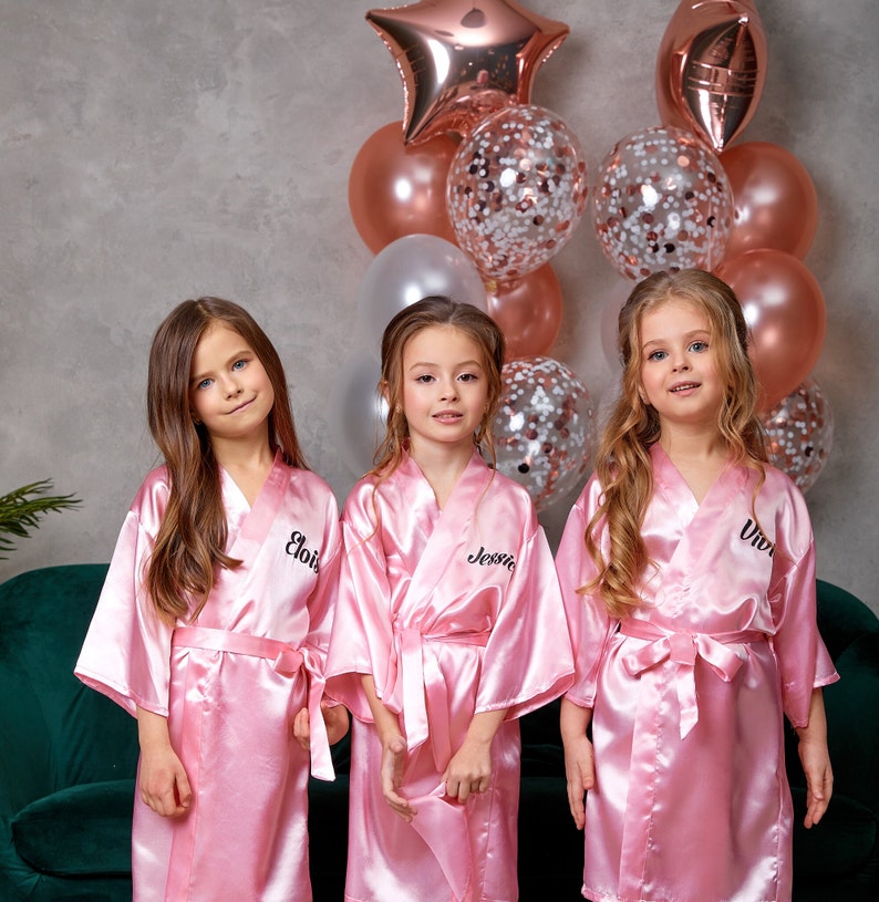 May include: Three young girls wearing matching pink satin robes with personalized names embroidered on the front. The robes are tied at the waist with a matching sash. Behind them are balloons in shades of pink, rose gold, and silver, including a star-shaped balloon.