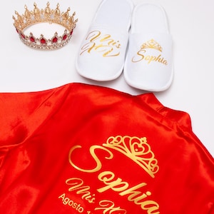 a red satin robe with gold text and a crown, along with a pair of white slippers with gold text and a crown.