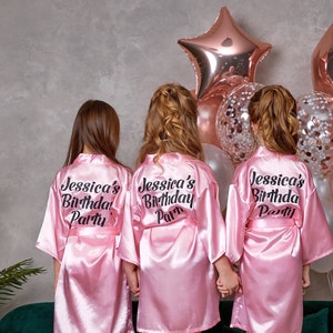 May include: Three children wearing pink satin robes with the text "Jessica's Birthday Party" in black. The robes have a matching belt. Behind them are rose gold star and round balloons, and balloons filled with confetti.