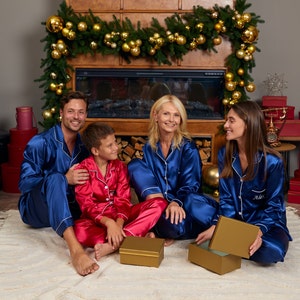 May include: A family of four wearing matching blue satin pajamas with white piping. They are sitting in front of a fireplace decorated with a garland of green pine branches and gold ornaments. The family is holding gold gift boxes.
