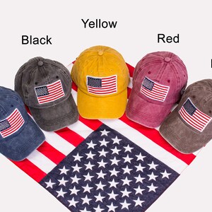 May include: Six baseball caps in different colors with an American flag patch on the front. The caps are arranged in a row on a red, white, and blue American flag bandana. The colors of the caps are navy blue, black, yellow, red, brown, and brown.