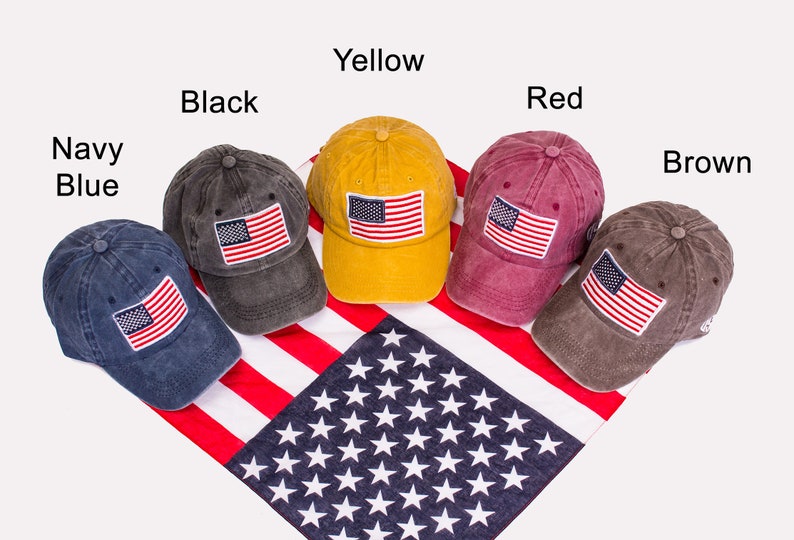 May include: Six baseball caps in different colors with an American flag patch on the front. The caps are arranged in a row on a red, white, and blue American flag bandana. The colors of the caps are navy blue, black, yellow, red, brown, and brown.