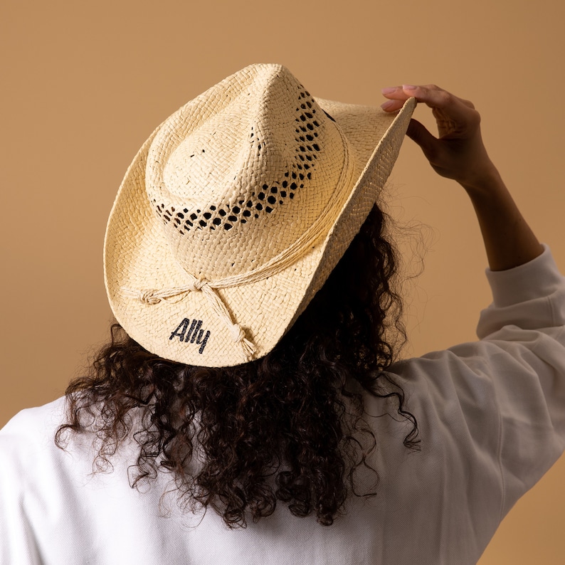 May include: A natural straw cowboy hat with a woven design and the word "Ally" printed in black. The hat has a decorative rope band and is being held up by a person.
