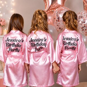 May include: Three girls wearing pink satin robes with the text "Jessica's Birthday Party" on the back. The robes are knee-length and have a matching belt. The background includes balloons and floral decorations.
