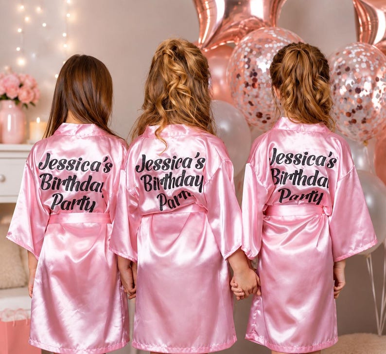 May include: Three girls wearing pink satin robes with the text "Jessica's Birthday Party" on the back. The robes are knee-length and have a matching belt. The background includes balloons and floral decorations.