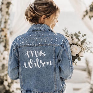 May include: A light blue denim jacket with pearl embellishments and the text "Mrs. Wilson" in white script. The date "06.07.2025" is printed on the upper back. The jacket is part of a bridal ensemble.