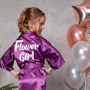 May include: A purple satin robe with the words "Flower Girl" in white script on the back. The robe is tied at the waist and worn by a young person. Balloons in shades of pink and white are in the background.