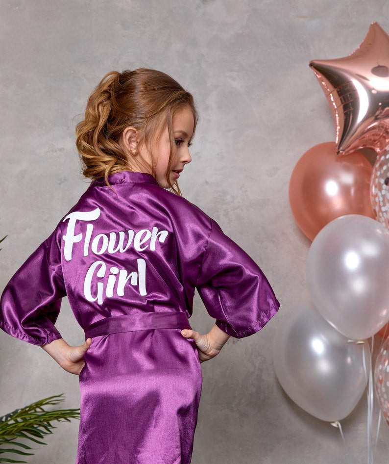 May include: A purple satin robe with the words "Flower Girl" in white script on the back. The robe is tied at the waist and worn by a young person. Balloons in shades of pink and white are in the background.