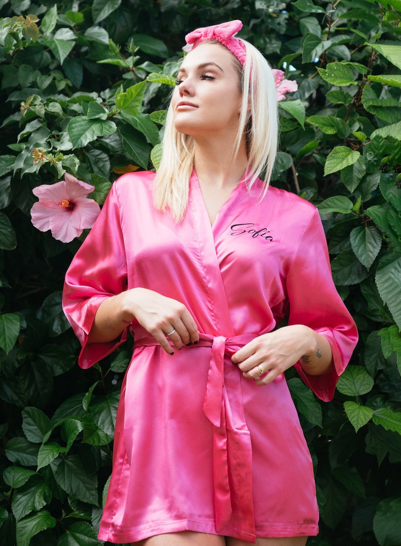 May include: A pink satin robe with a black embroidered name "Sofia" on the left side. The robe has a matching pink sash and is tied at the waist.