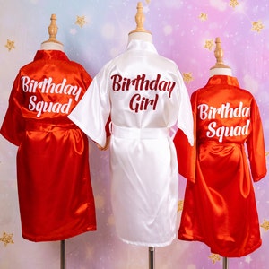 May include: Three satin robes on mannequins. Two are red with "Birthday Squad" in white script, and one is white with "Birthday Girl" in red script. The robes have a shiny finish and are tied with matching belts.