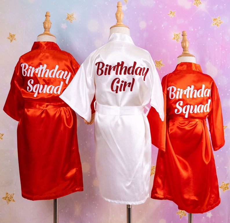 May include: Three satin robes on mannequins. Two are red with "Birthday Squad" in white script, and one is white with "Birthday Girl" in red script. The robes have a shiny finish and are tied with matching belts.