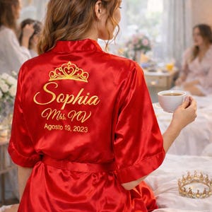 A woman wearing a red robe with the name "Sophia" and the date "Ages 19, 2023" printed on it, sitting on a bed with a cup of coffee in her hand.