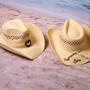 May include: Two straw cowboy hats with a light beige color. One hat has a decorative heart-shaped accent, and the other hat has the name "Lara" printed on it. Both hats have a woven design and a brim.