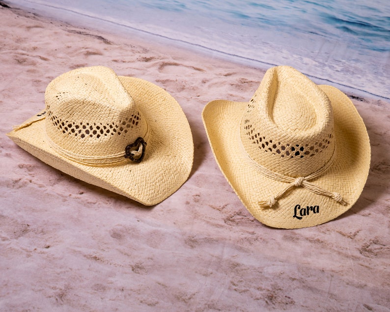 May include: Two straw cowboy hats with a light beige color. One hat has a decorative heart-shaped accent, and the other hat has the name "Lara" printed on it. Both hats have a woven design and a brim.