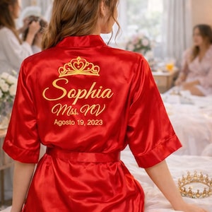 A woman wearing a red satin robe with the name "Sophia" and the date "Agosto 19, 2023" printed on it, sitting on a bed with a crown and other personal items.