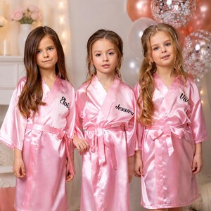 May include: Three young girls wearing matching pink satin robes with personalized names embroidered on the front. The robes have a wrap-around design with a matching belt. Balloons are visible in the background.