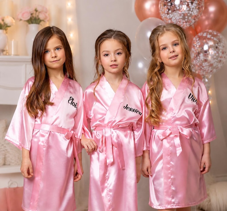 May include: Three young girls wearing matching pink satin robes with personalized names embroidered on the front. The robes have a wrap-around design with a matching belt. Balloons are visible in the background.
