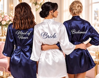 May include: Three satin robes with tie waists and three-quarter length sleeves. The centre robe is white with the word Bride embroidered in blue. The two outer robes are navy blue with the words Maid of Honor and Bridesmaid in white.