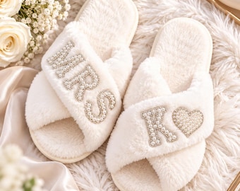 May include: A pair of white plush open-toe slippers with crisscross straps. The left slipper features the letters MRS in white faux pearls and rhinestones. The right slipper features the letter K and a heart shape in white faux pearls and rhinestones.