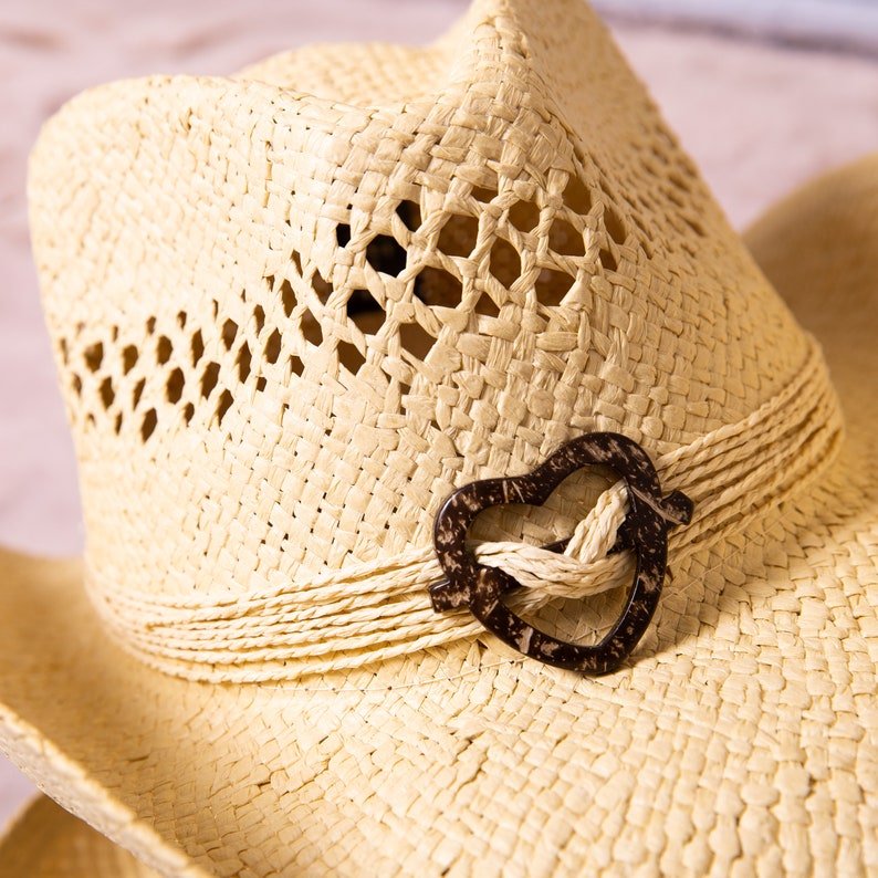 May include: A straw cowboy hat with a heart-shaped accent. The hat features a woven design with decorative cutouts and a brown heart-shaped embellishment. The brim is wide, and the hat is a light beige color.