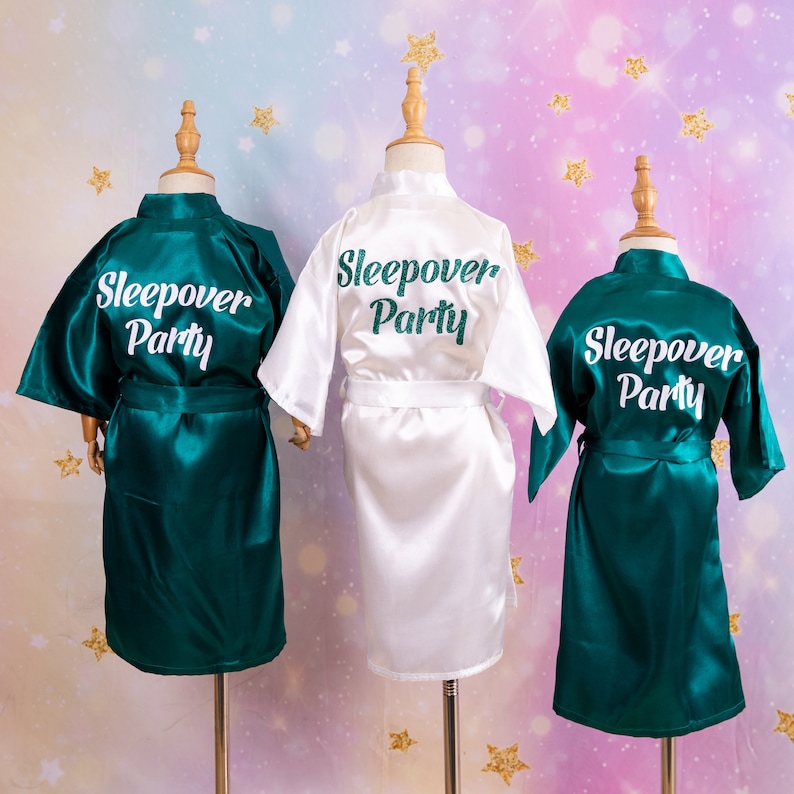 May include: Three satin robes on display. Two are emerald green, and one is white. Each robe has the words "Sleepover Party" embroidered on the back in a matching color. The robes are displayed on wooden mannequin forms.