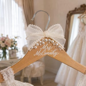May include: A light-colored wooden clothes hanger featuring a large, sheer white organza bow at the top. The hanger is decorated with a cluster of white pearls and displays the white script text "Mrs. McConahy" printed on the wooden surface.