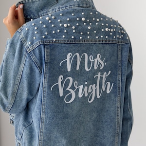 May include: A blue denim jacket with pearl embellishments on the back. The jacket has the text "Mrs. Brighth" and the date "10.28.18" embroidered on the back.
