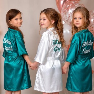 May include: Three young people wearing satin robes. Two robes are teal and say "Birthday Squad" in white script on the back. The third robe is white and says "Birthday Girl" in green script. Balloons are in the background.